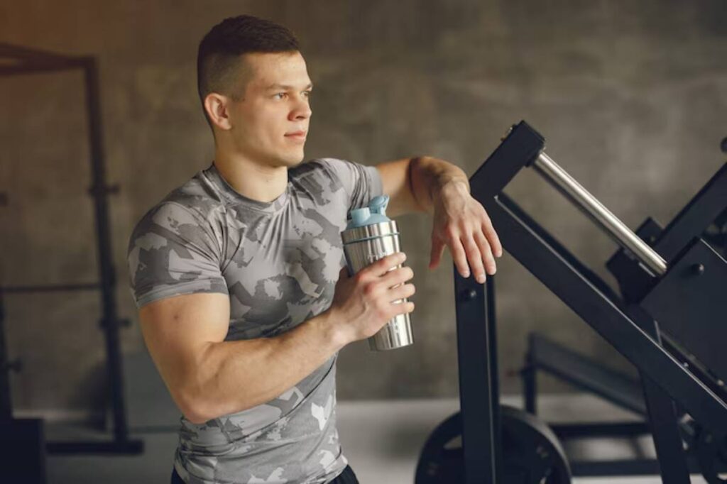 A muscular man in a gray camo workout shirt is holding a shaker bottle while leaning against gym equipment.