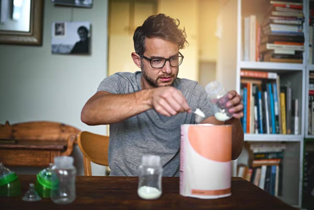 A person in a gray shirt measures formula powder into a container, surrounded by baby bottles on a wooden table, with books in the background.