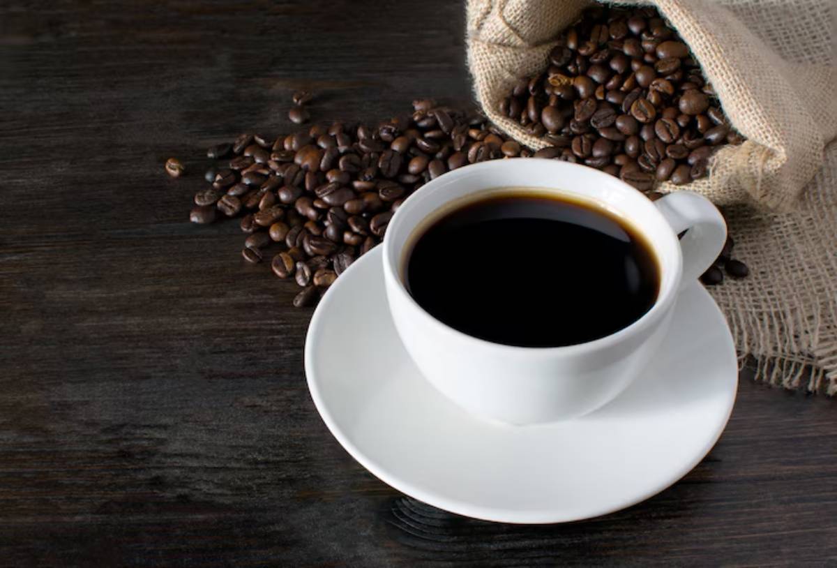 A white cup of black coffee sits on a plate next to spilled coffee beans and a burlap sack on a dark wooden surface.