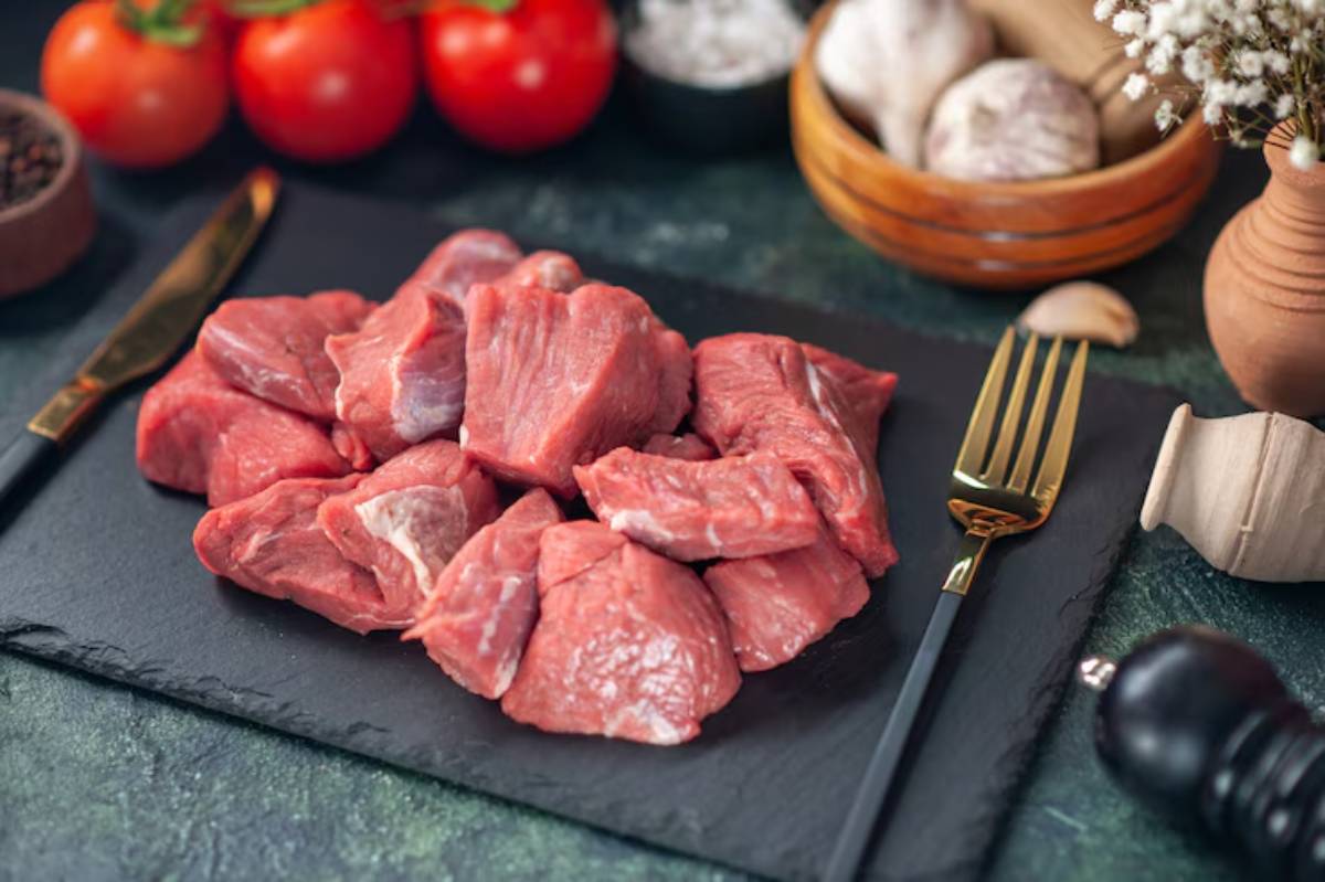Fresh chunks of raw red meat on a slate platter, surrounded by tomatoes, garlic, and kitchen utensils, ready for cooking.