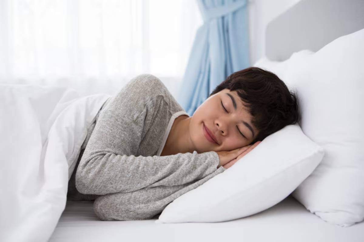 A person in a cozy gray sweater lies on a bed, resting their head on a white pillow, surrounded by soft bedding and natural light.
