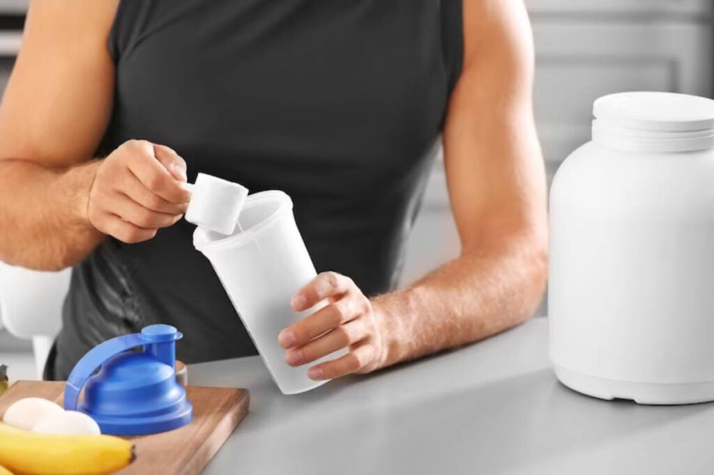 A person in a sleeveless shirt adds a scoop of protein powder into a clear container while bananas and eggs rest on the table.