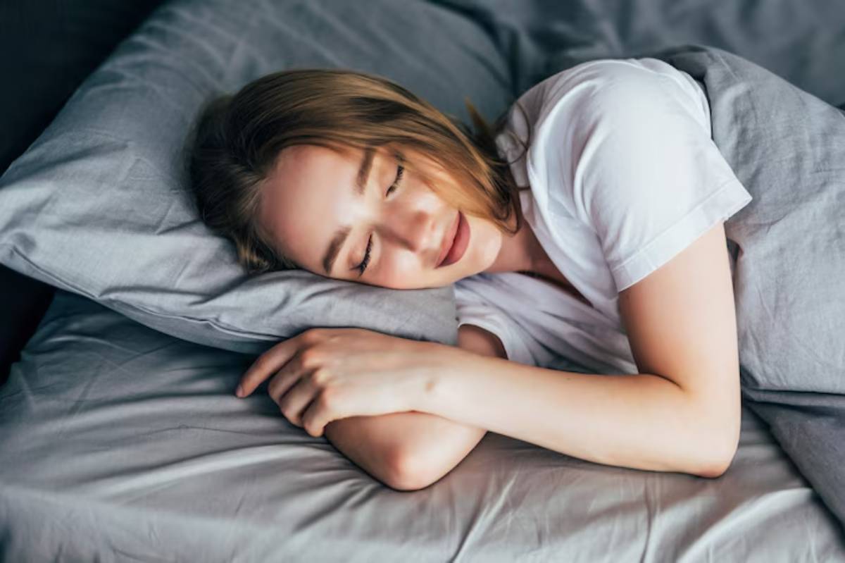 A person lies on gray bedding, resting their head on a pillow, with arms wrapped around themselves in a relaxed pose.