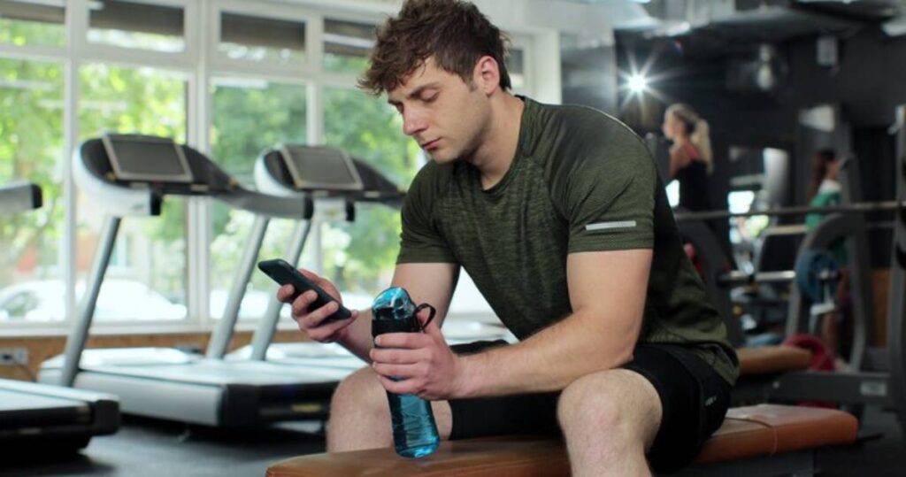 A person sits on a bench in a gym, looking at their phone while holding a water bottle, surrounded by exercise equipment and natural light.