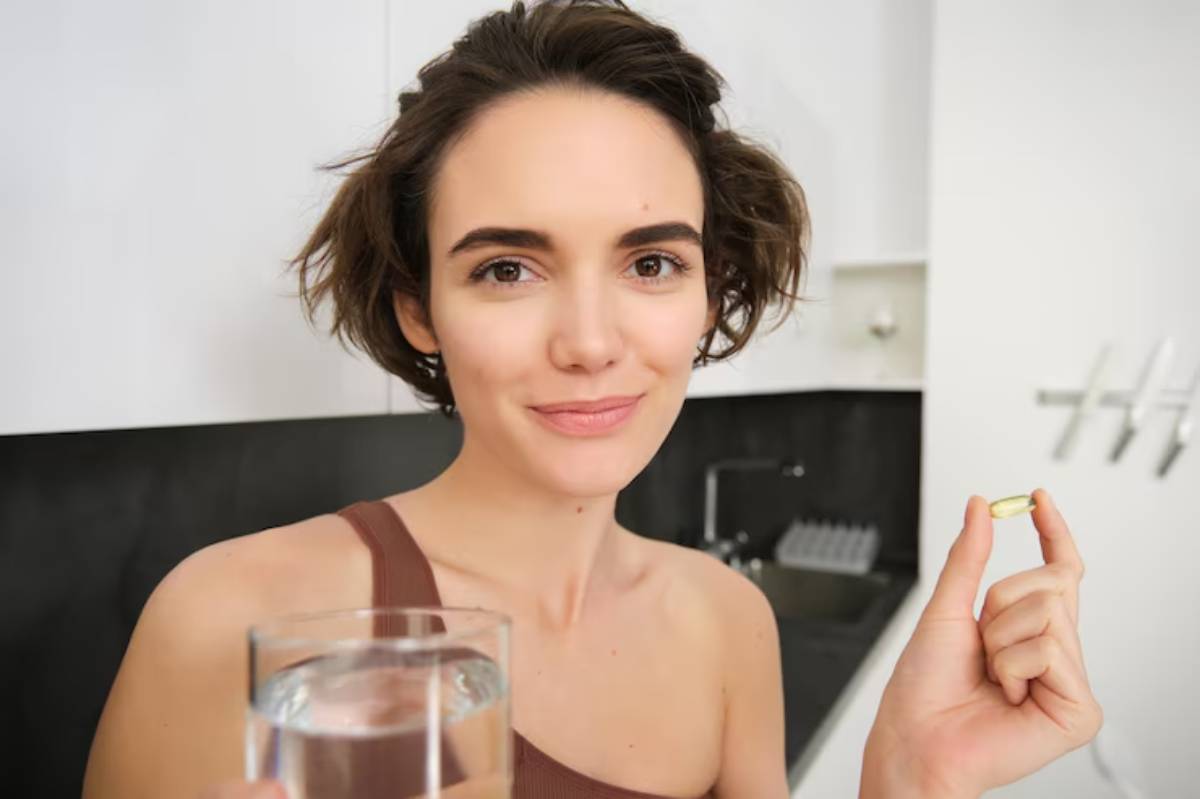 A person holds a glass of water in one hand and a capsule in the other, standing in a well-lit kitchen.