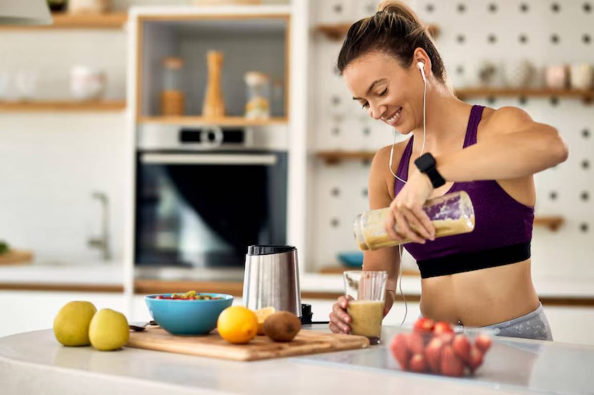 A woman in a fitness outfit prepares a smoothie in a modern kitchen, surrounded by fresh fruits and a blender on the countertop.
