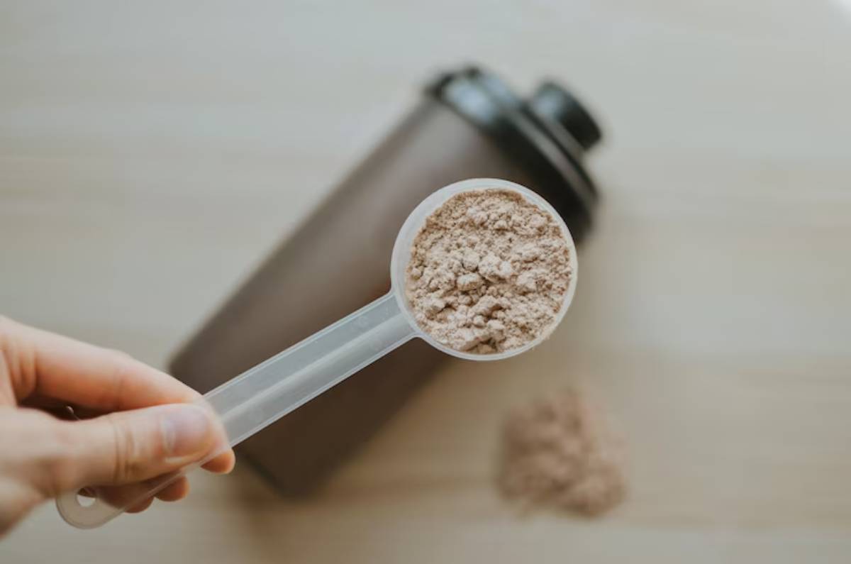 A hand holds a clear scoop of powder above a dark shaker bottle, with loose powder spilled nearby on a wooden surface.