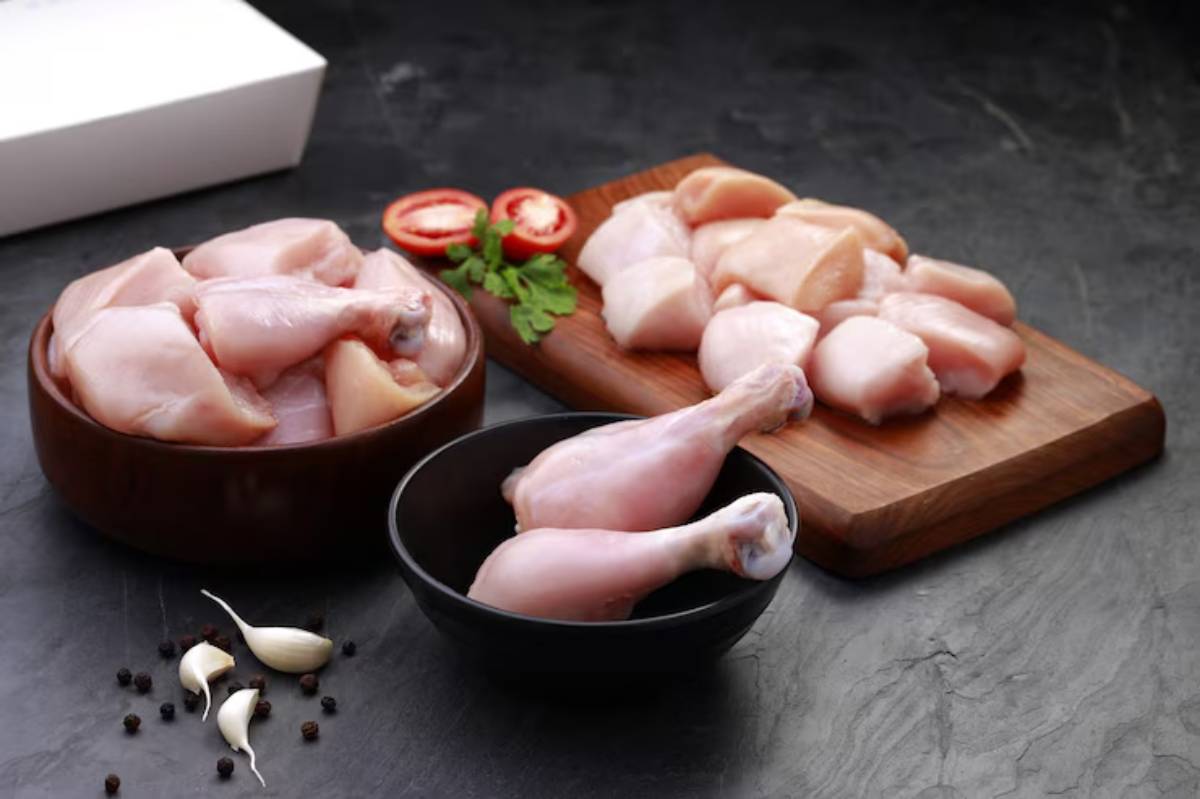 Raw chicken pieces arranged in two bowls and on a cutting board, with fresh tomatoes and garlic nearby on a dark surface.