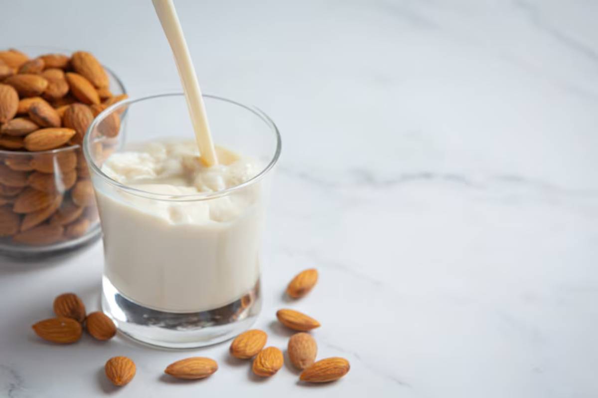 A glass of creamy almond milk is being poured, surrounded by whole almonds on a marble surface, showcasing a healthy beverage option.