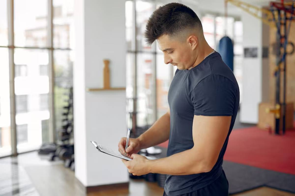 A person in a gym wearing a black shirt takes notes on a clipboard, surrounded by exercise equipment and large windows.