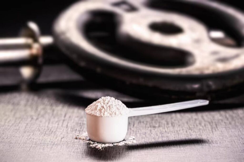 A white scoop of protein powder sits on a dark surface, with a weight plate blurred in the background, suggesting fitness and nutrition.