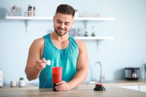 A person in a teal tank top adds a scoop to a red shaker bottle on a wooden kitchen counter, surrounded by small indoor plants.