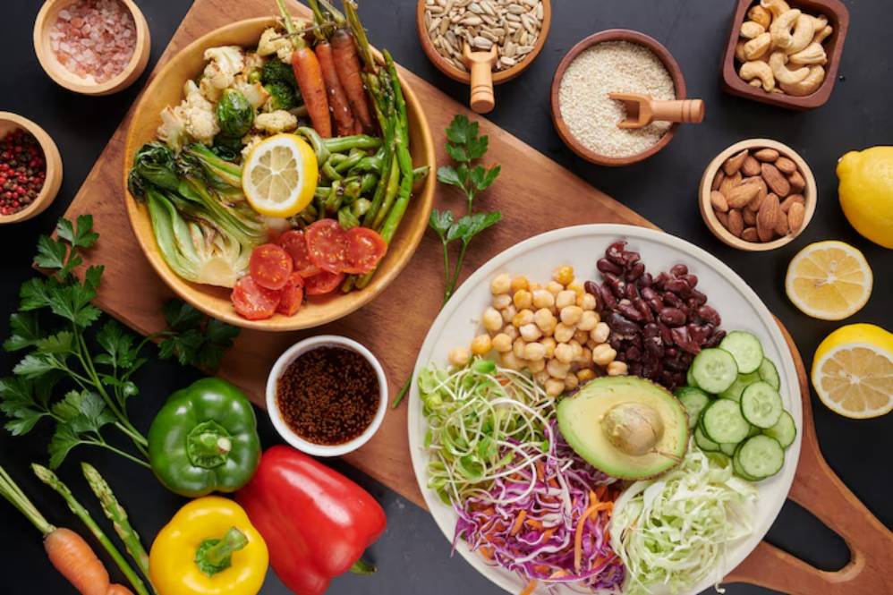 A vibrant assortment of fresh vegetables, legumes, nuts, and seeds arranged on a wooden board, with lemon slices and herbs.