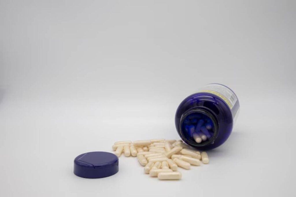 A blue bottle spills white capsules onto a light gray surface, with the bottle cap nearby.