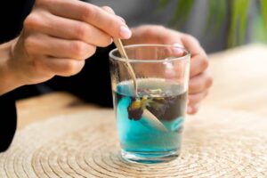 A person uses a spoon to stir a glass of vibrant blue liquid with a flower submerged inside, set on a textured surface.