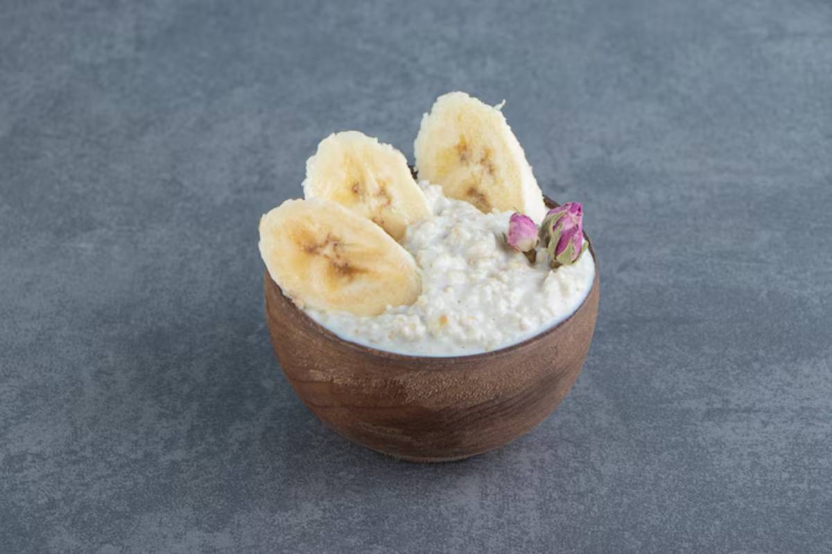 A wooden bowl filled with creamy oatmeal topped with banana slices and pink rosebud accents on a gray surface.