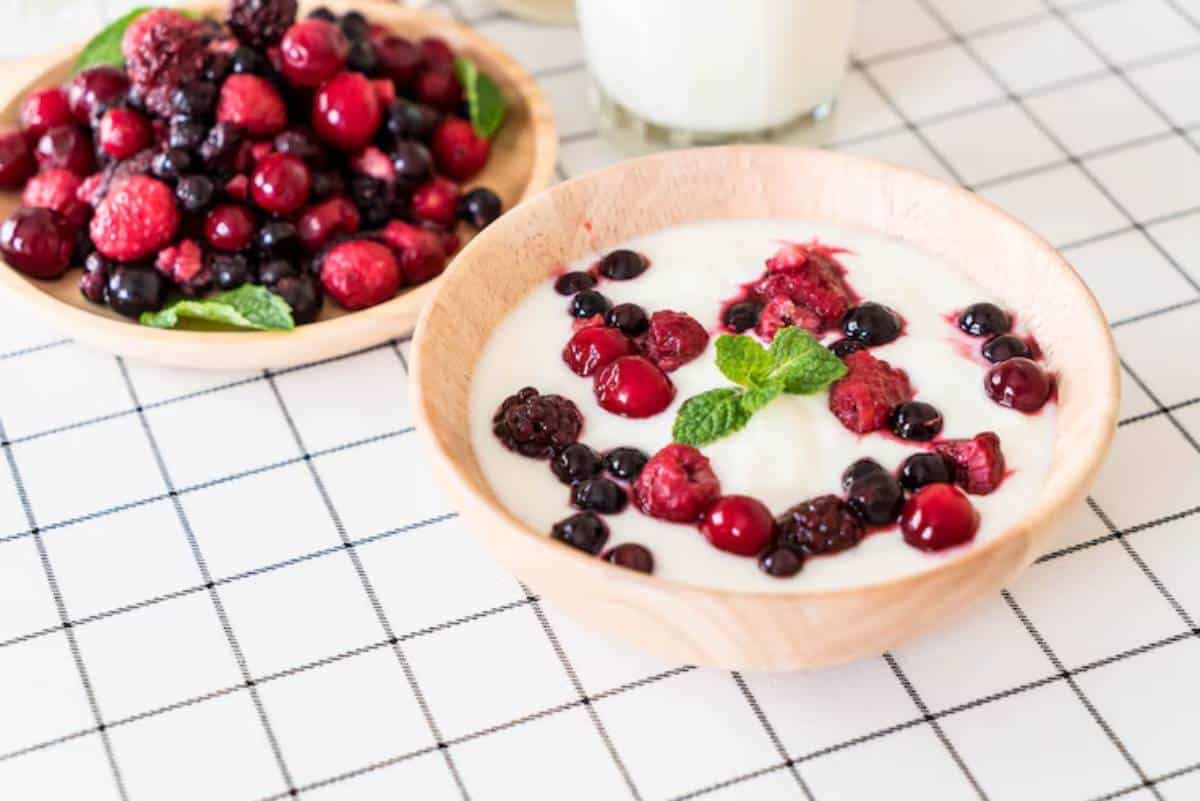 A wooden bowl filled with yogurt topped with various berries and a mint leaf, alongside a plate of fresh mixed berries.