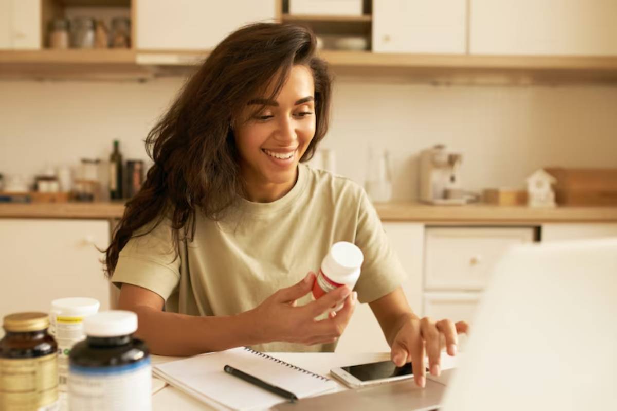 A person sits at a table surrounded by various bottles; they hold a pill bottle while using a phone and notebook.
