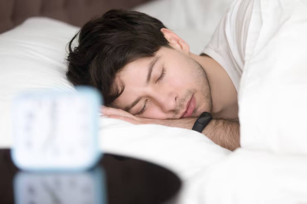 A person sleeps comfortably in bed, partially curled under white blankets, with a small blue alarm clock on a dark bedside table.